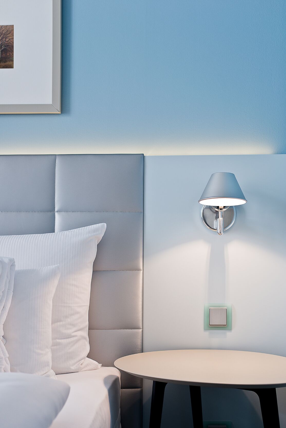 Ein modernes Hotelzimmer mit einer gepolsterten Kopfteil, einem Nachttisch, einer Lampe und einem gerahmten Bild an der Wand.
