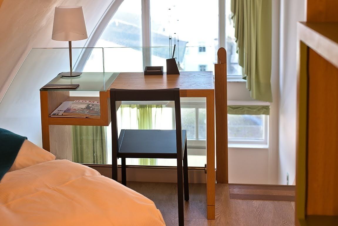 A bedroom with a bed, a desk with a lamp, a chair, and a window with curtains.