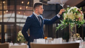 Un hombre con traje azul sostiene una copa de vino en una mesa con centros de flores.