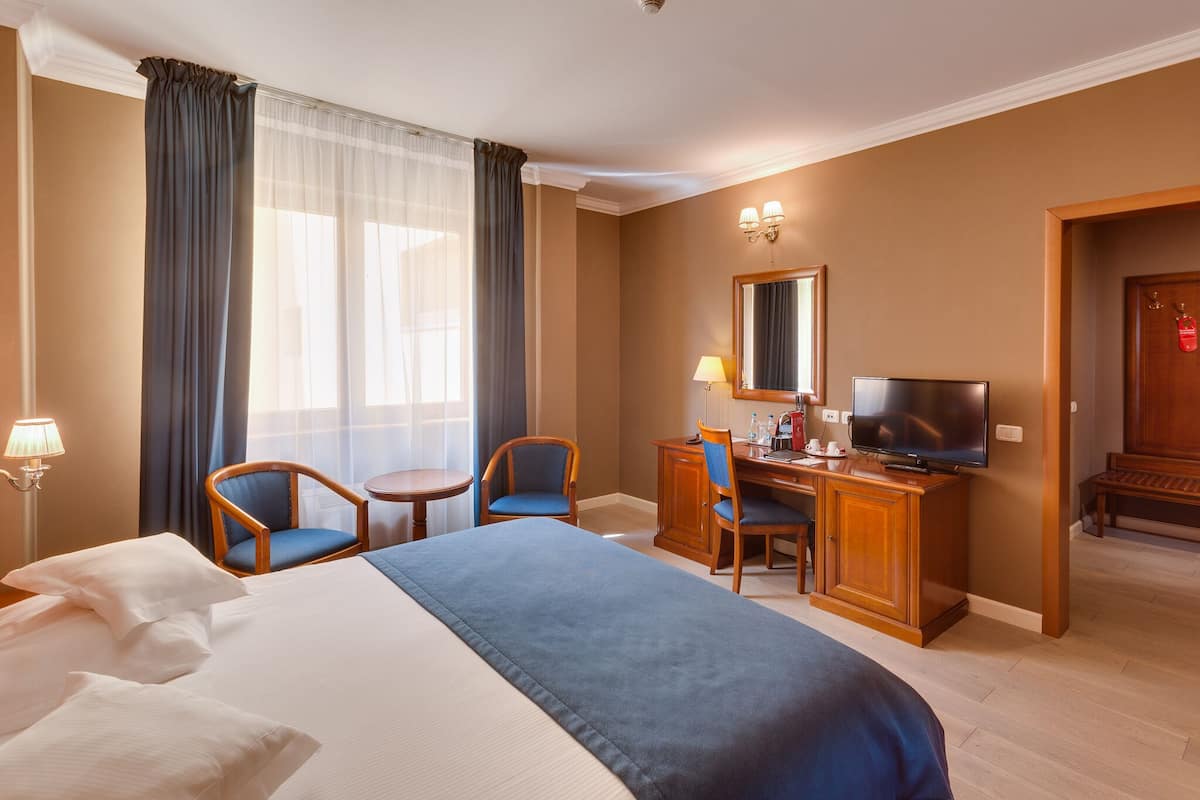 A hotel room with a bed, two chairs, a desk with a television, and a mirror.
