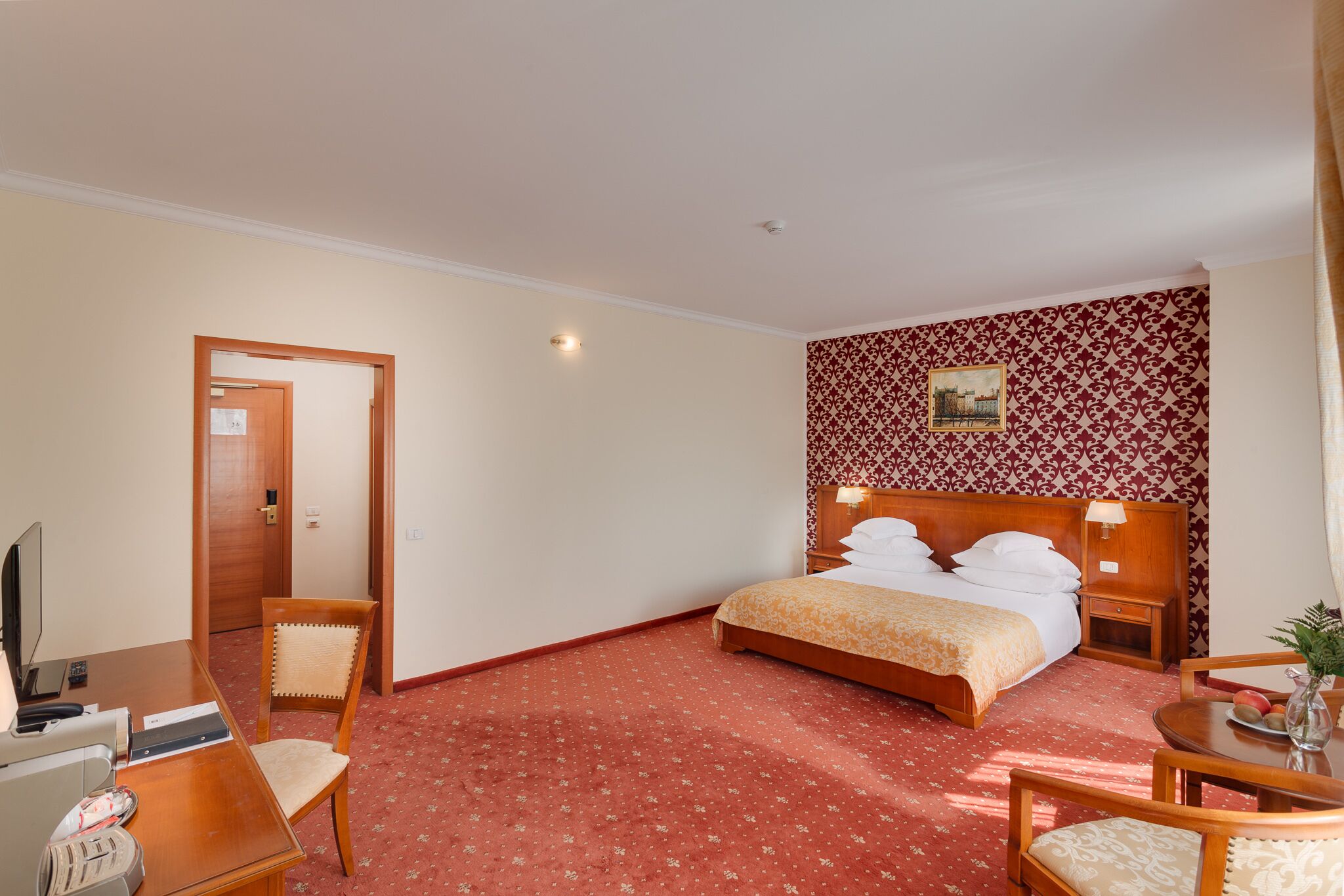 Habitació d'hotel amb un llit, un escriptori amb un ordinador, una cadira, una taula petita amb un vas, i una paret amb un disseny decoratiu.