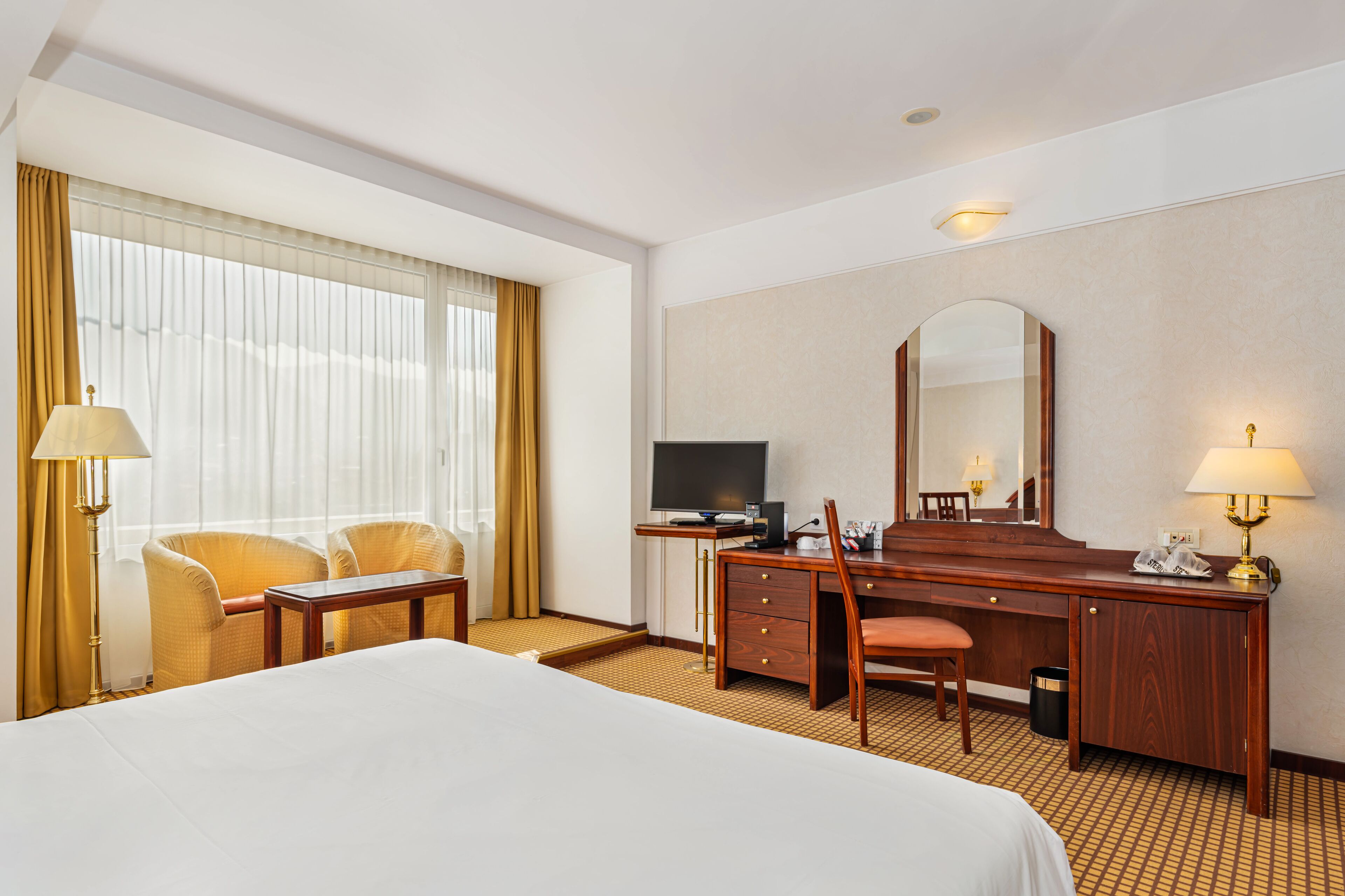 A hotel room with a bed, a desk with a computer, a chair, a TV, and a large mirror.
