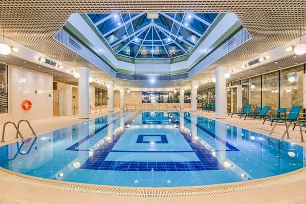 Una amplia piscina interior con techo de cristal, rodeada de sillones de descanso y suelo de baldosas.