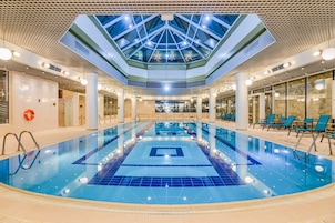 Una amplia piscina interior con techo de cristal, rodeada de sillones de descanso y suelo de baldosas.
