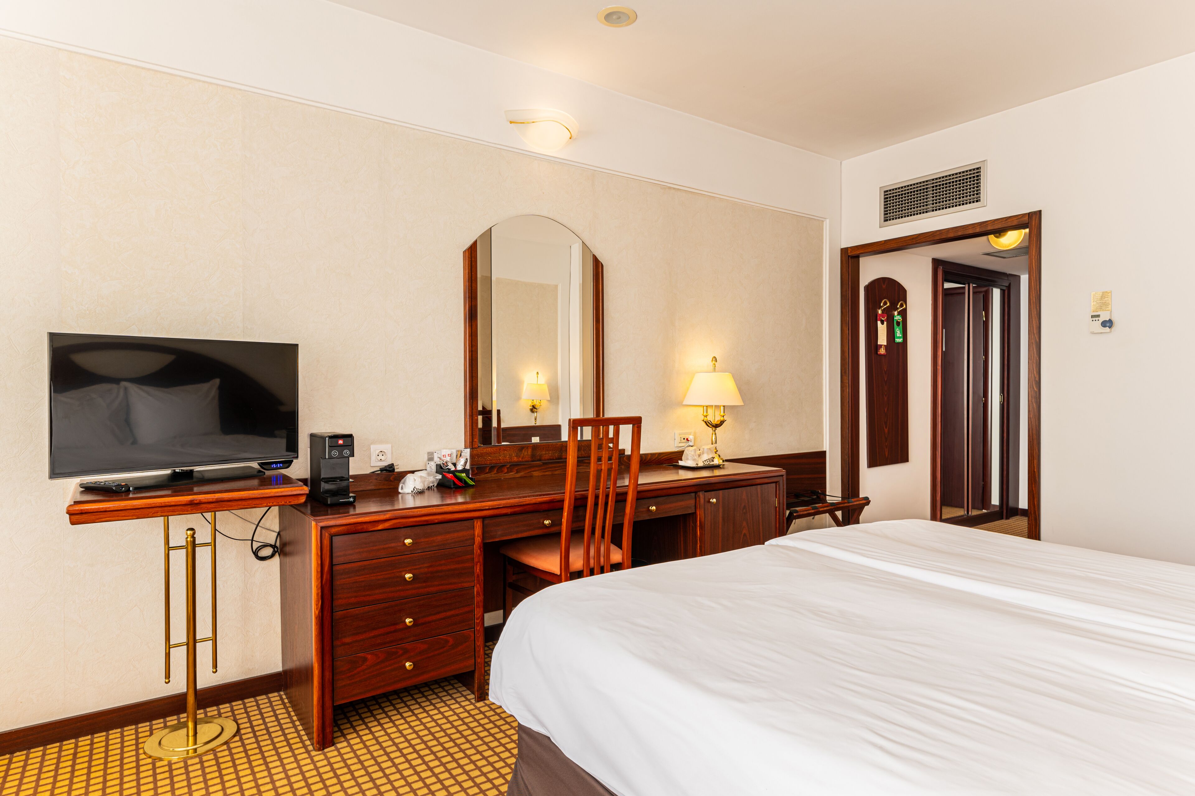 A hotel room with a bed, a desk with a TV, a chair, a lamp, and a mirror.