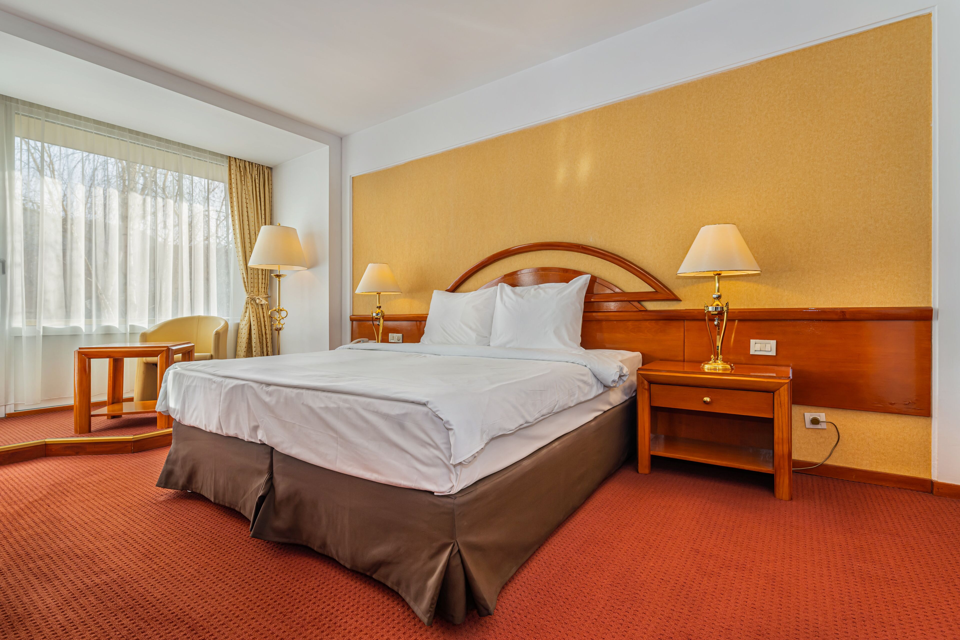 A hotel room with a large bed, two bedside tables with lamps, a wooden headboard, and a view of trees through sheer curtains.
