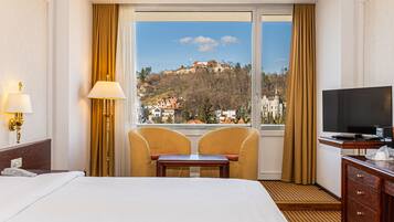 A hotel room with a bed, a desk, two chairs, a television, and a large window offering a view of a hill with buildings.