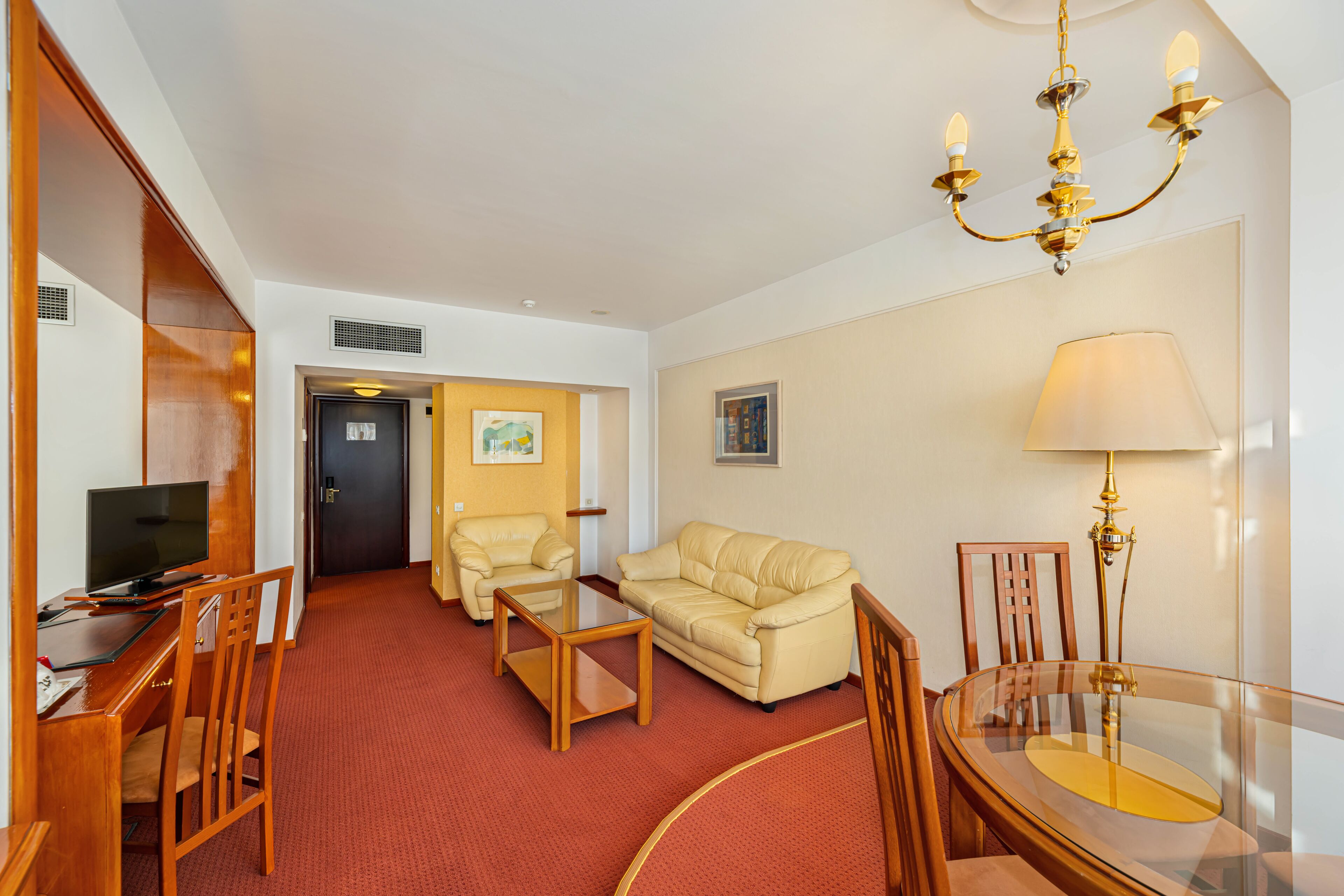 Habitación de hotel con alfombra roja, sofá color beige, una silla de madera, una mesa de centro de cristal, un televisor sobre un escritorio de madera, una lámpara de araña y una lámpara.