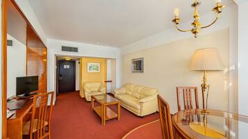 Habitación de hotel con alfombra roja, sofá color beige, una silla de madera, una mesa de centro de cristal, un televisor sobre un escritorio de madera, una lámpara de araña y una lámpara.