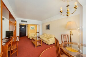 Habitación de hotel con alfombra roja, sofá color beige, una silla de madera, una mesa de centro de cristal, un televisor sobre un escritorio de madera, una lámpara de araña y una lámpara.