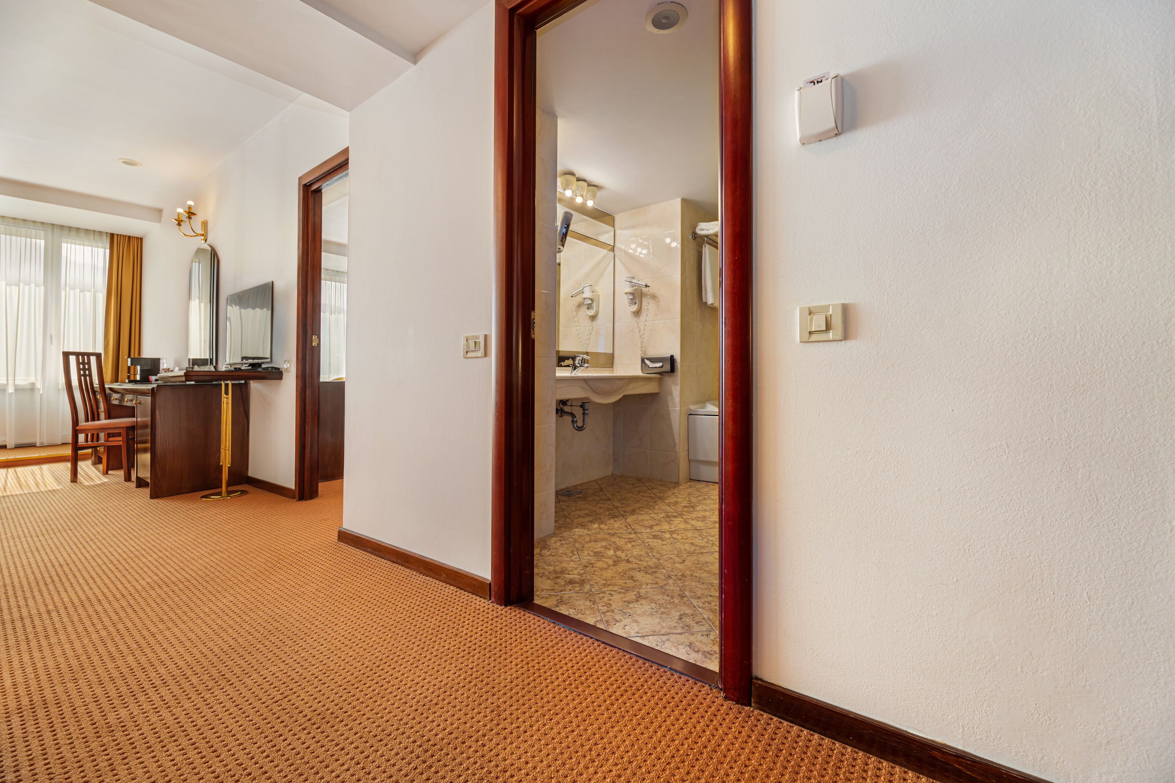 Habitación de hotel con piso de madera, un escritorio con silla, un espejo grande y un baño visible a través de una puerta abierta.