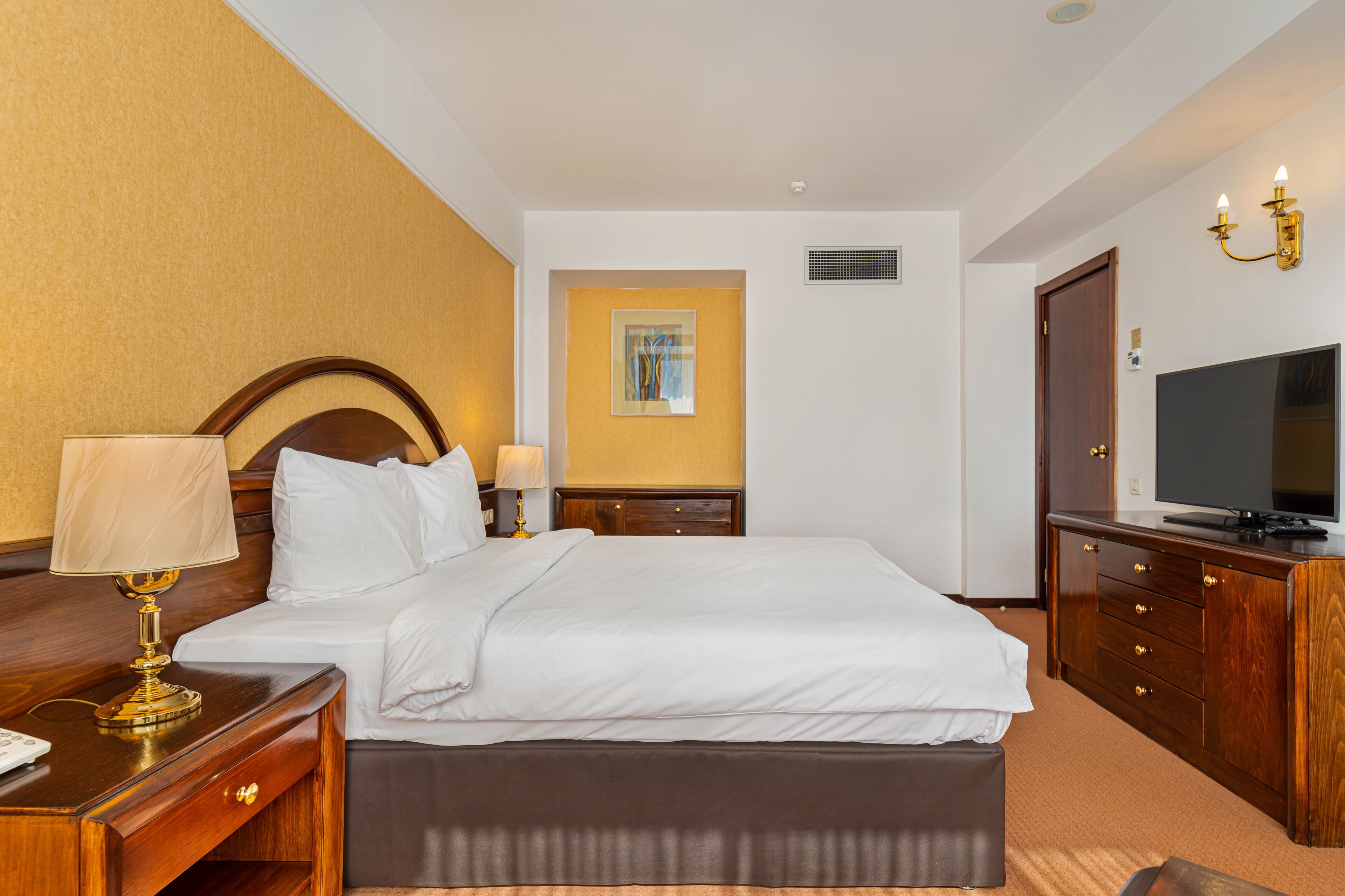 A hotel room with a large bed, a bedside table with a lamp, a dresser with a flat-screen TV, and a painting on the wall.