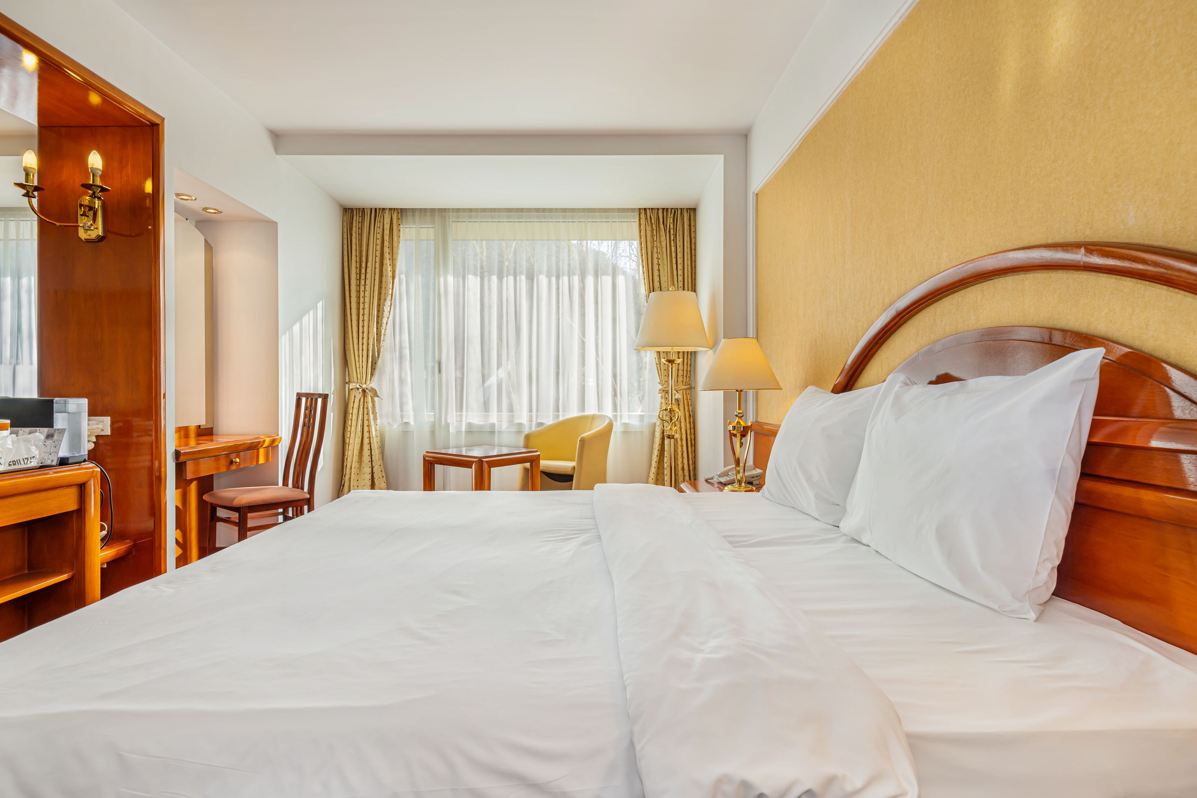 A hotel room with a large bed, a desk with a chair, a lamp, and a window with curtains.