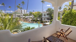 A balcony with wooden chairs and a table overlooking a pool area with lounge chairs and umbrellas, surrounded by palm trees and buildings.
