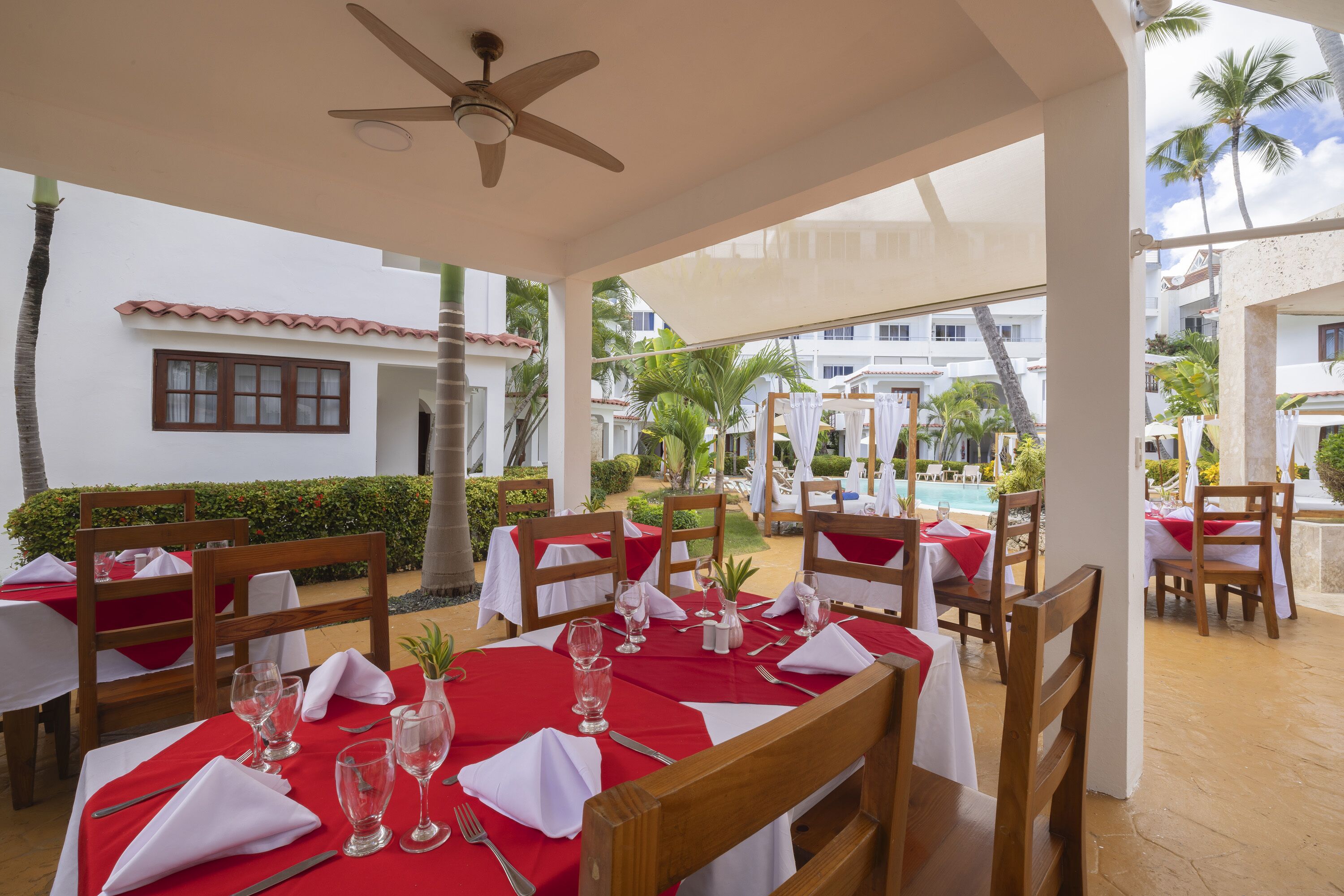 An outdoor dining area with tables set for two, wooden chairs, and a ceiling fan.