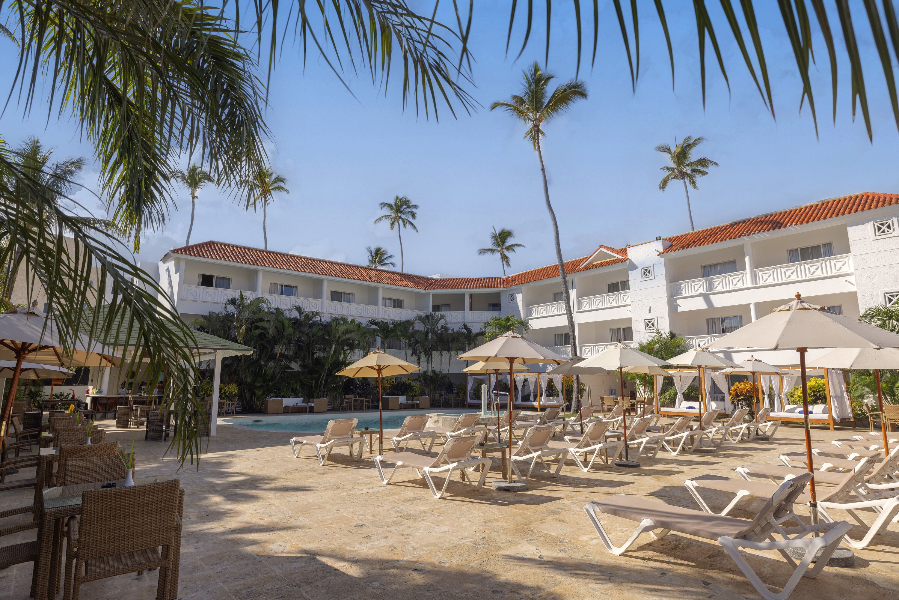 A resort pool area with sun loungers and umbrellas, surrounded by palm trees and a building with a tiled roof.