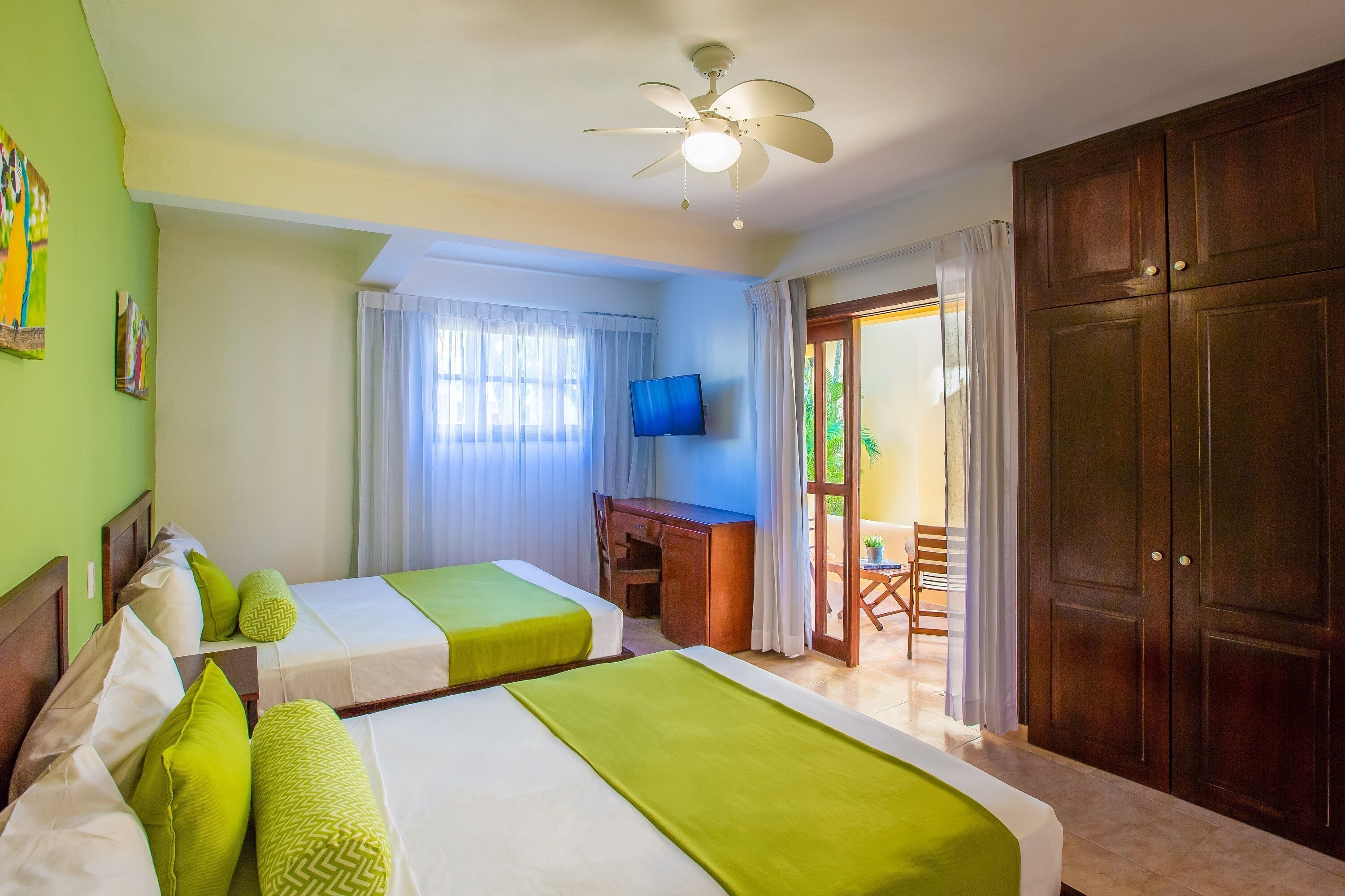 A hotel room with two beds, a desk, a television, and a balcony with a chair.