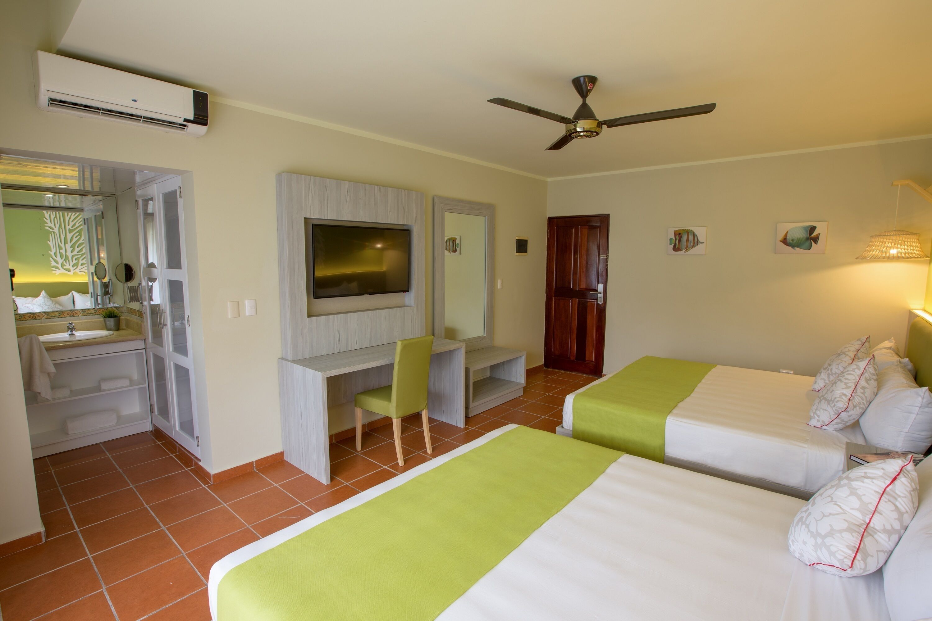 A hotel room with two beds, a desk, a television, a ceiling fan, and a bathroom with a sink and mirror.