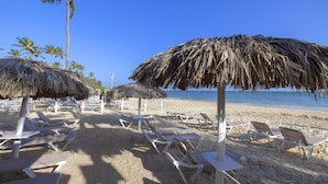 A beach with rows of sun loungers and thatched umbrellas, palm trees, and a clear blue sky.