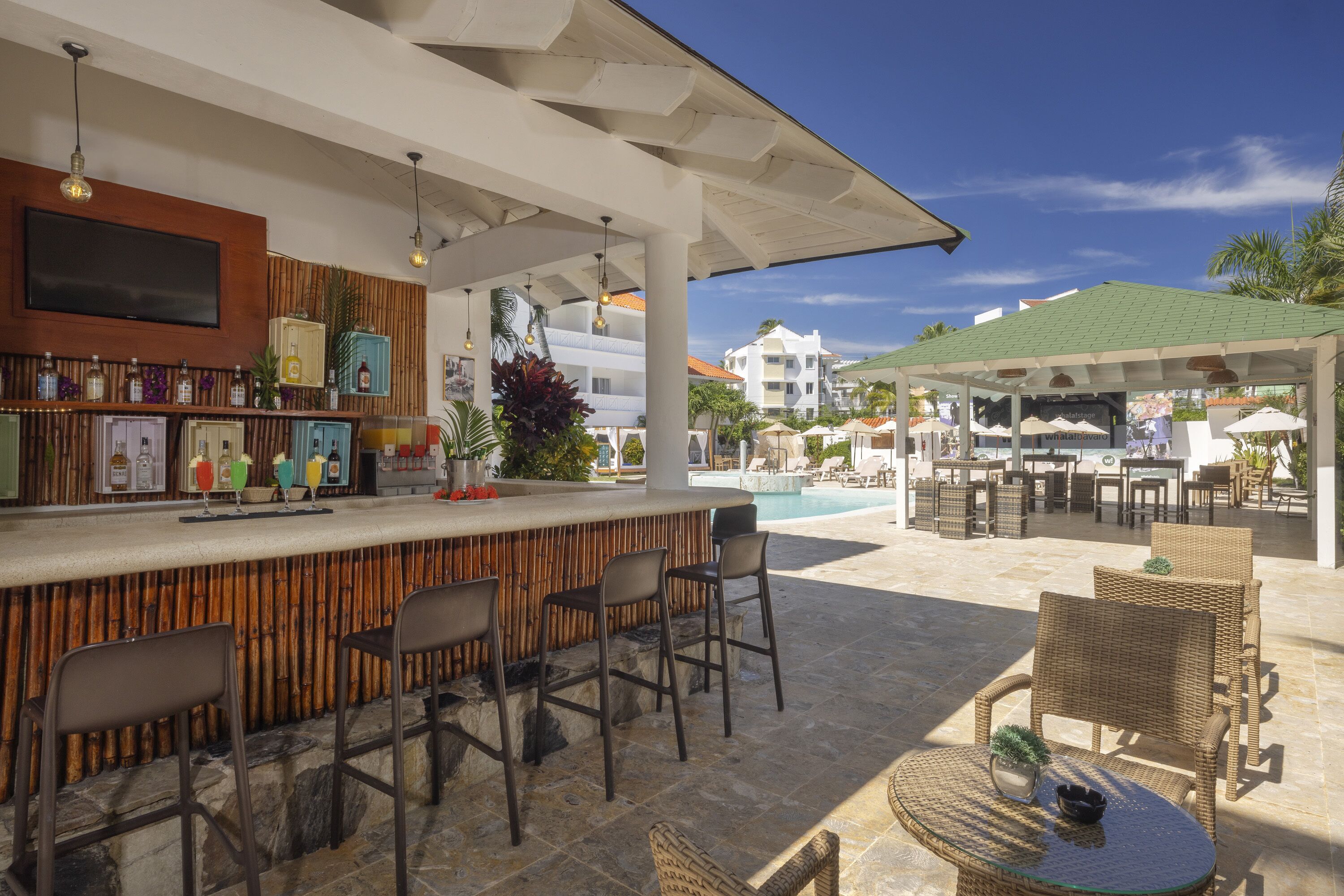 An outdoor bar area with a poolside seating arrangement, a bar counter with various bottles, and a view of the pool and buildings in the background.