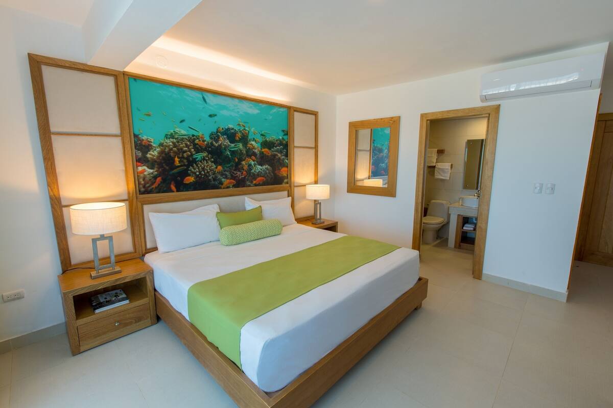 A hotel room with a large bed, a nightstand, a mirror, a bathroom, and a painting of an underwater scene.