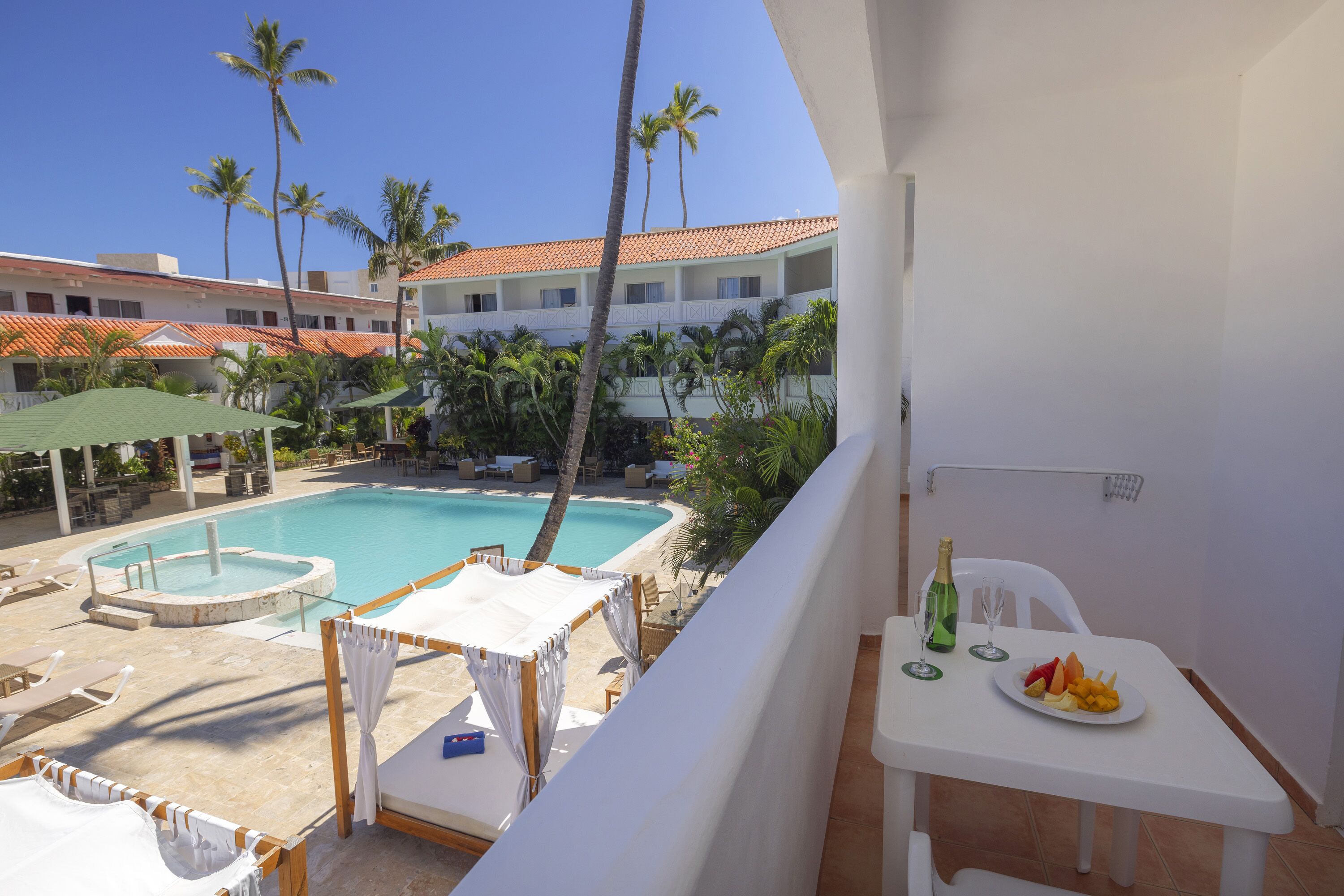 A balcony with a pool, palm trees, and a dining table with fruit and drinks.