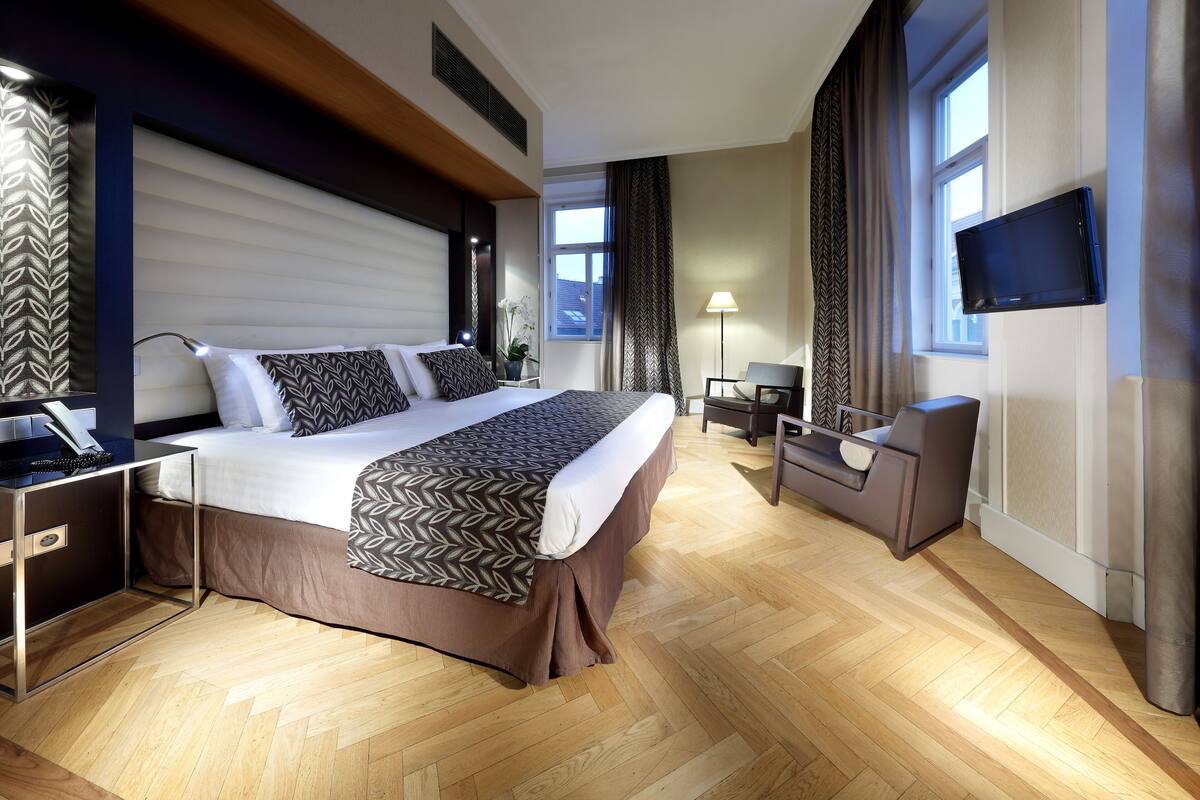A modern hotel room with a large bed, bedside tables, a desk, a chair, a TV, and a window with curtains.