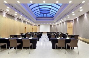 Uma sala de conferências com teto alto, iluminação embutida e claraboia. Mesas dispostas para uma reunião, com cadeiras e garrafas de água.
