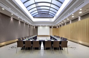 Sala de conferência com teto alto e claraboia, equipada com uma mesa comprida e cadeiras, e uma tela de projetor.