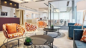 A modern hotel lobby with a reception desk, seating area with patterned chairs, a blue sofa, and a glass partition separating the dining area. - Eurostars Thalia (Prague)