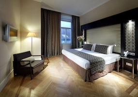 Quarto de hotel com cama, sofá, escrivaninha, luminária e televisão.
