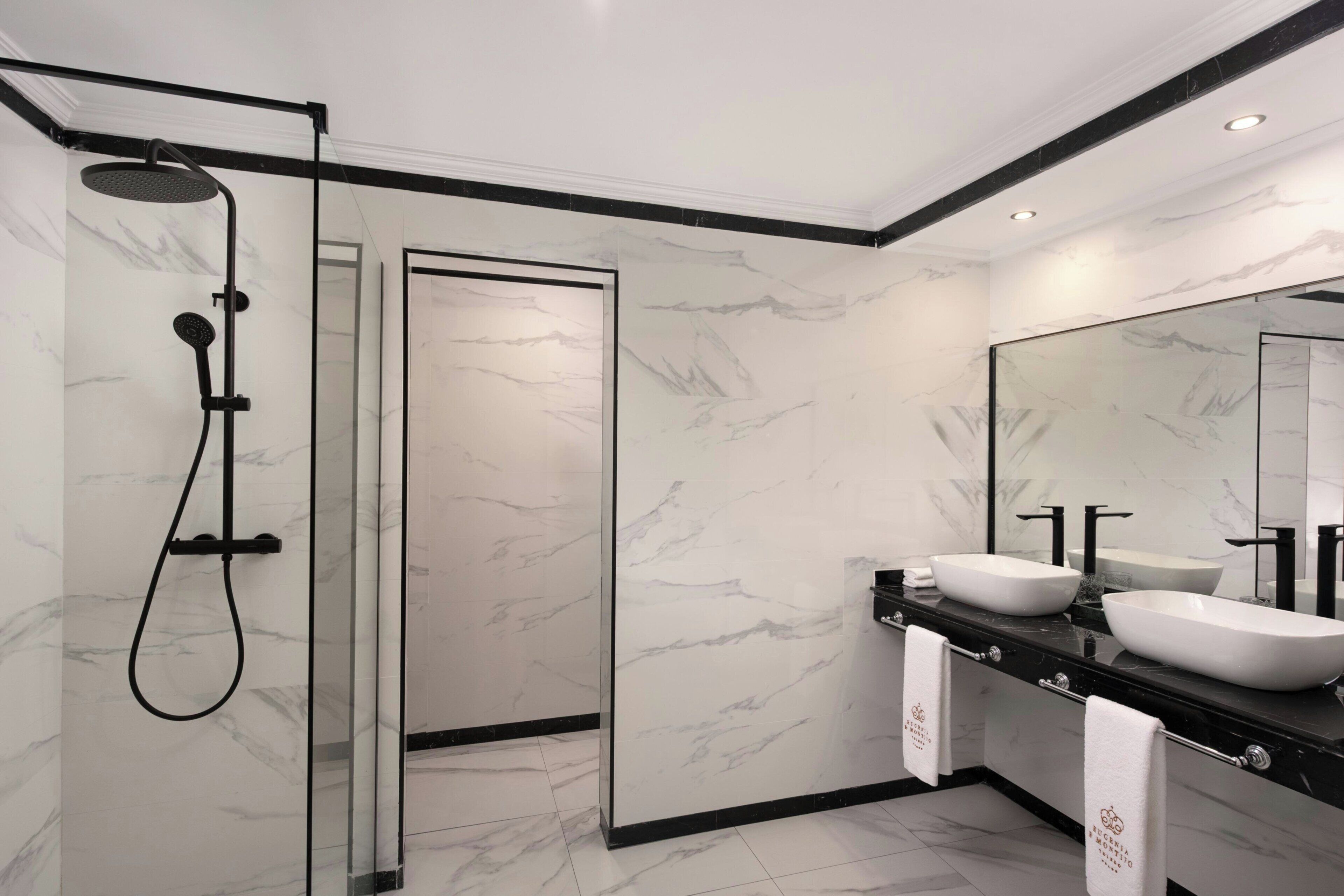 Ein modernes Badezimmer mit einer in Glas eingefassten Dusche, einer schwarzen Ablagefläche mit zwei Waschbecken und Wänden im Marmor-Look.