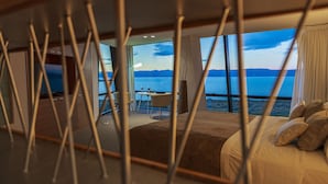 Una camera da letto con un'ampia finestra che si affaccia su un lago e montagne. Si vede un letto con i cuscini e un comodino con una lampada.