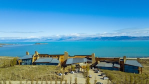 Un edificio moderno con una grande facciata vetrata e un tetto spiovente, affacciato su uno specchio d'acqua e montagne lontane.