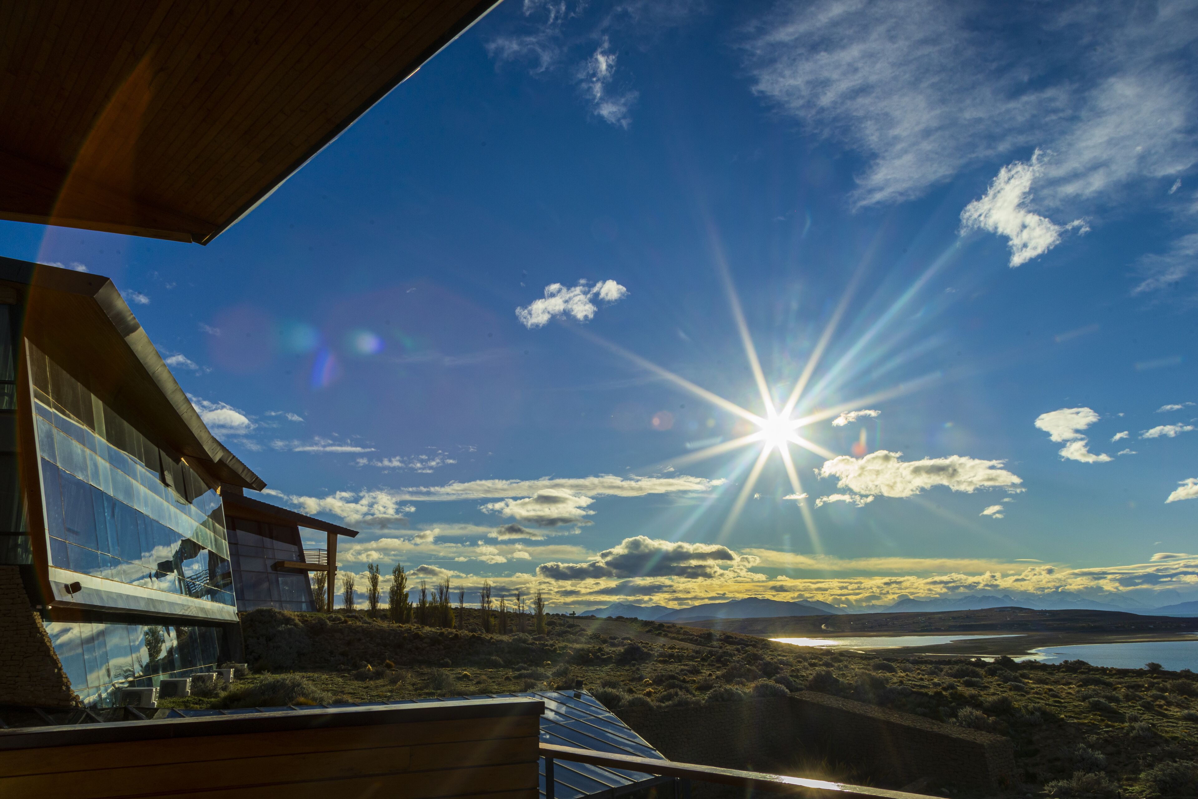 Ein modernes Gebäude mit großen Glasfenstern, einer Holzterrasse und Blick auf einen See und Berge.