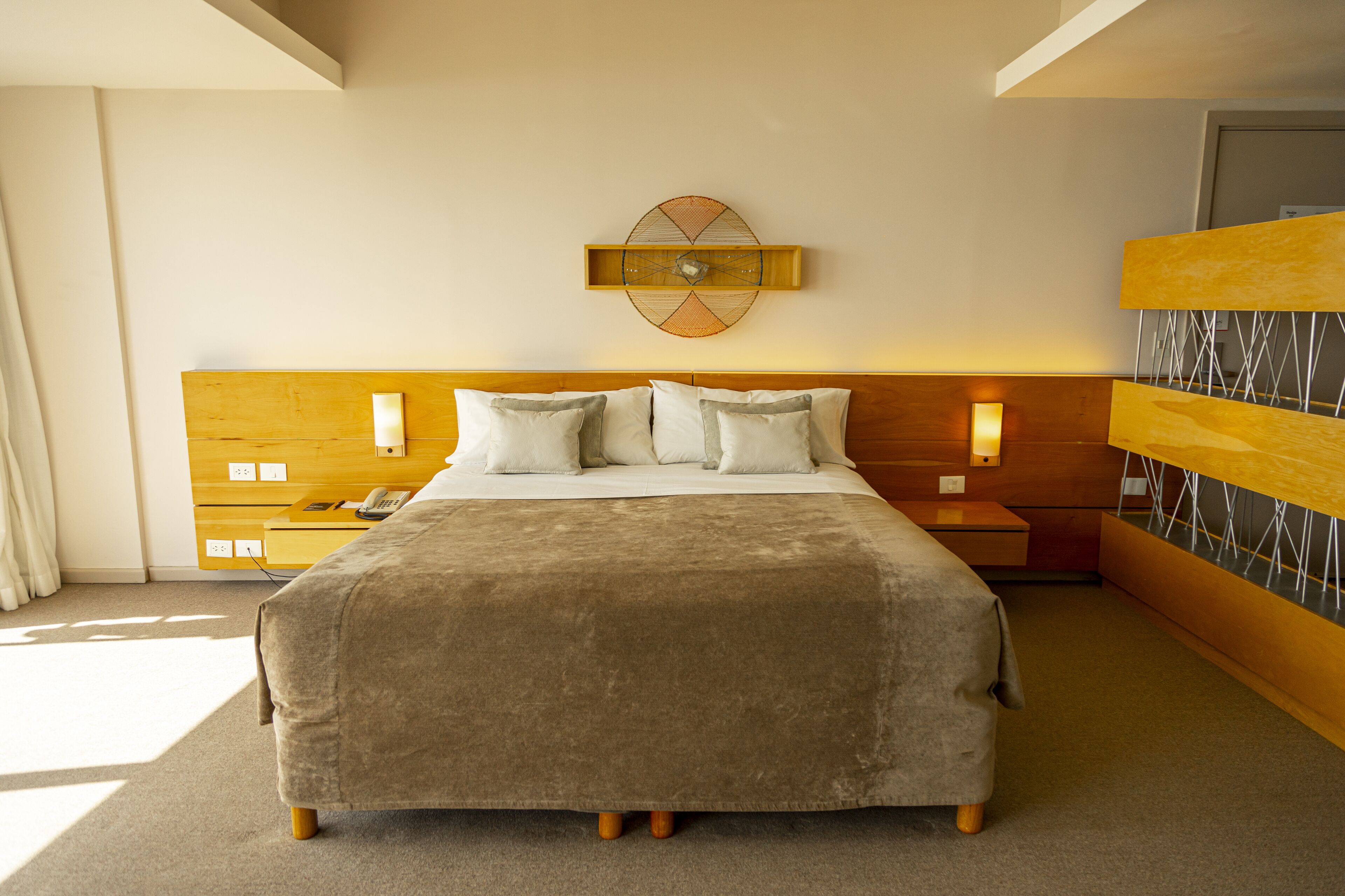 Quarto de hotel moderno com uma cama grande, cabeceira de madeira, relógio fixado na parede e mesa de cabeceira com luminária.