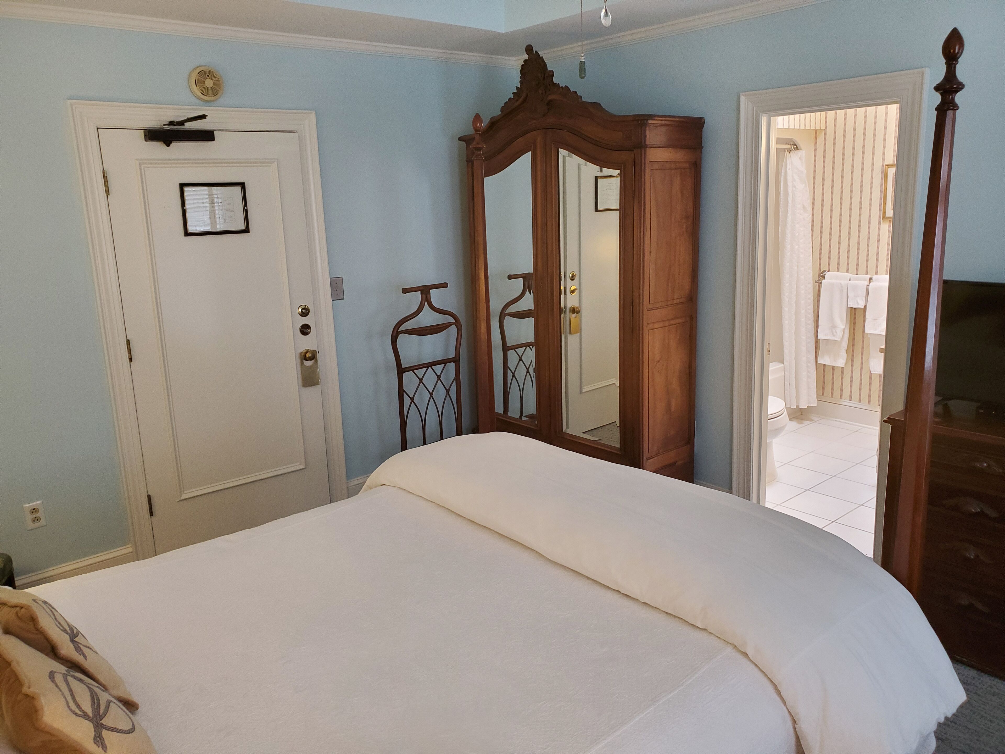 Een slaapkamer met een bed, een houten kledingkast, een spiegel en een deur met lichtschakelaar en slot.