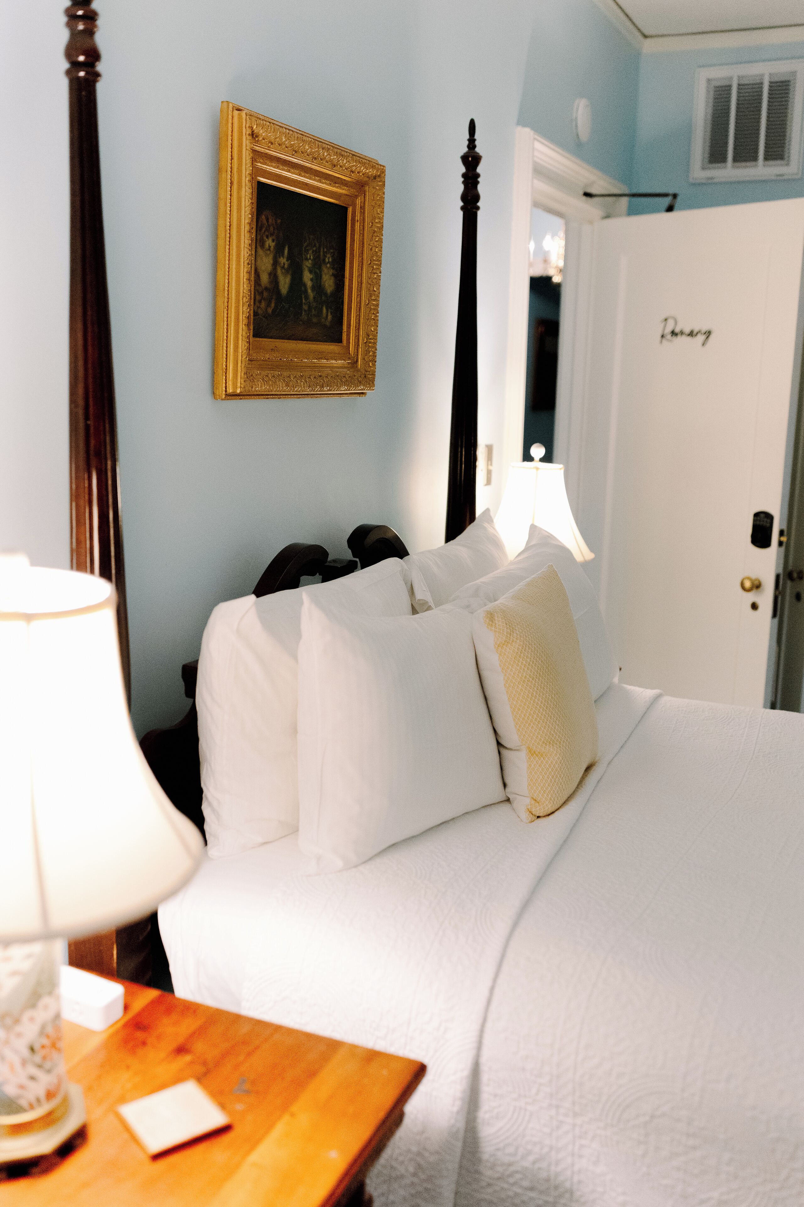 A bedroom with a four-poster bed, a bedside table with a lamp, and a framed picture on the wall.