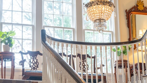 A grand staircase with white balusters, a chandelier, and large windows with a view of trees. - Morehead Inn (Charlotte)