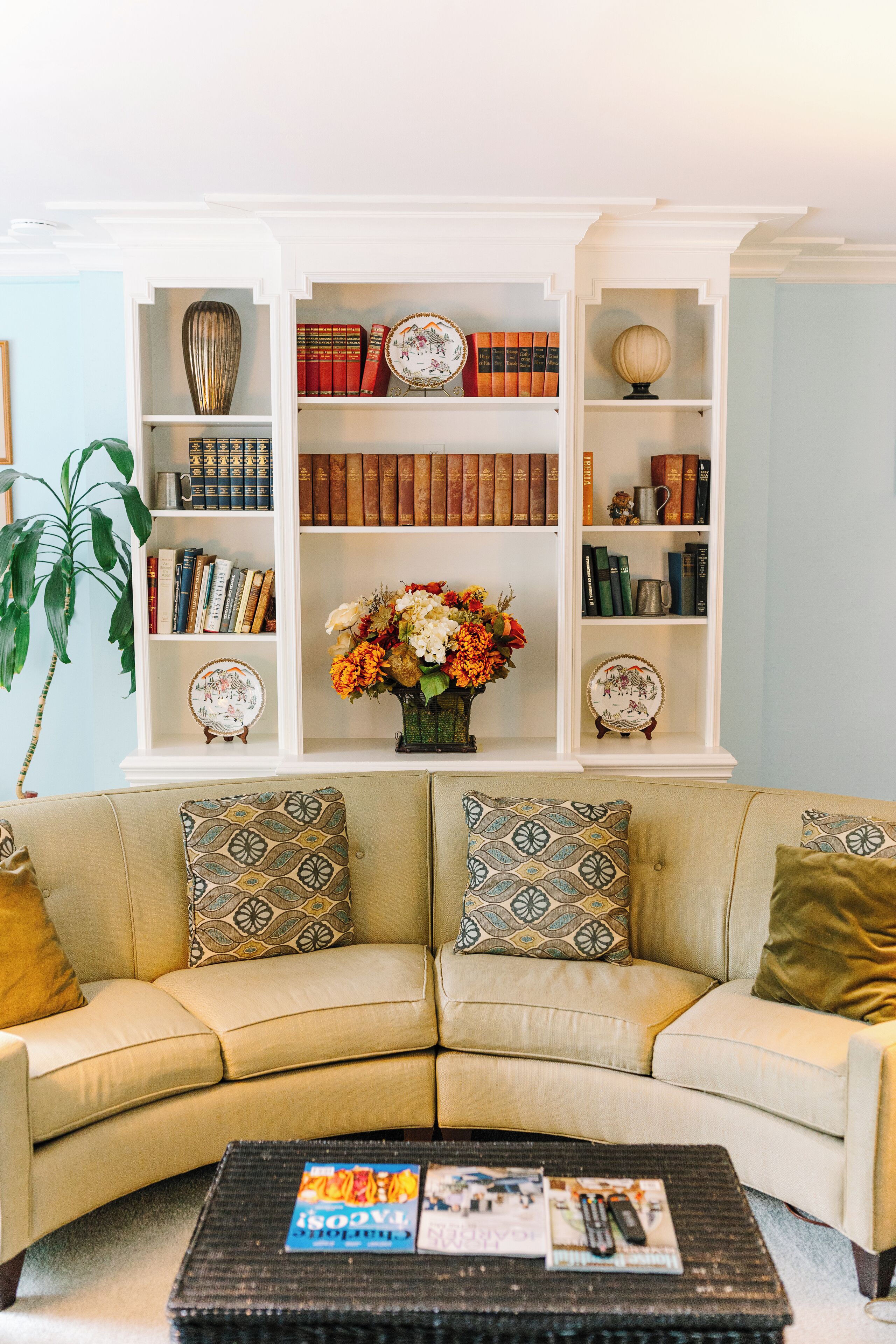 A living room with a beige sofa, patterned cushions, a coffee table with magazines and remote controls, and a bookshelf with books and decorative items.