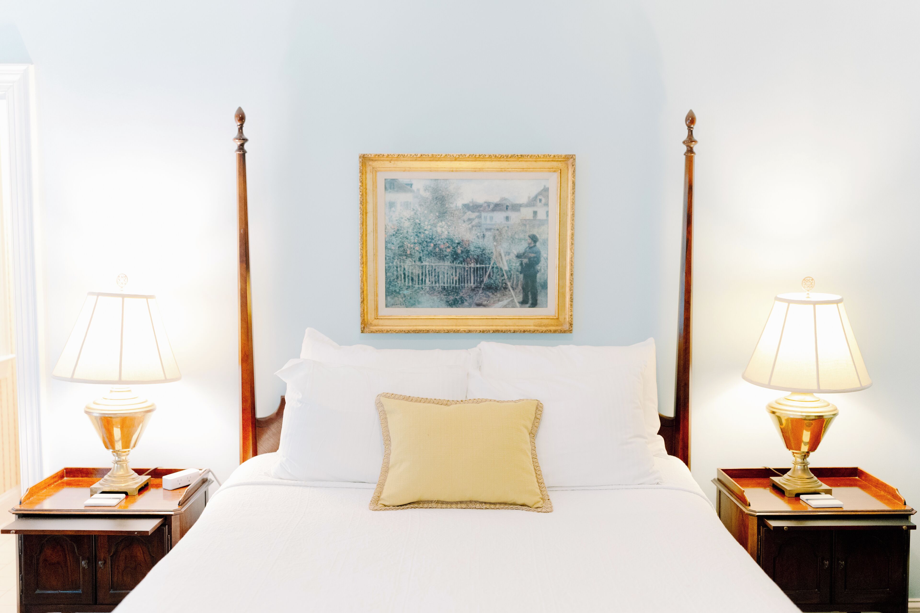 A neatly made bed with white linens, a single yellow pillow, and two bedside tables with lamps. A framed picture hangs above the bed.