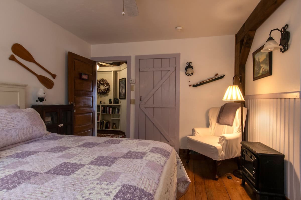 A bedroom with a large bed, a wooden wardrobe, a chair, a fireplace, and a bookshelf.