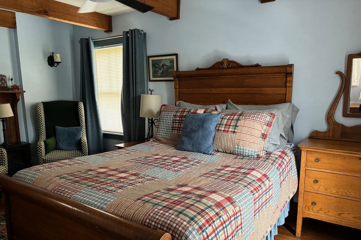 A bedroom with a wooden bed, a chair, a nightstand, a lamp, and a window with curtains.