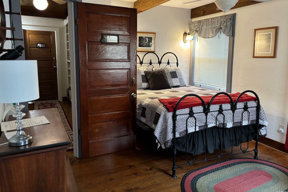 A bedroom with a wooden door, a bed with patterned bedding, a ceiling fan, and a window with blinds.