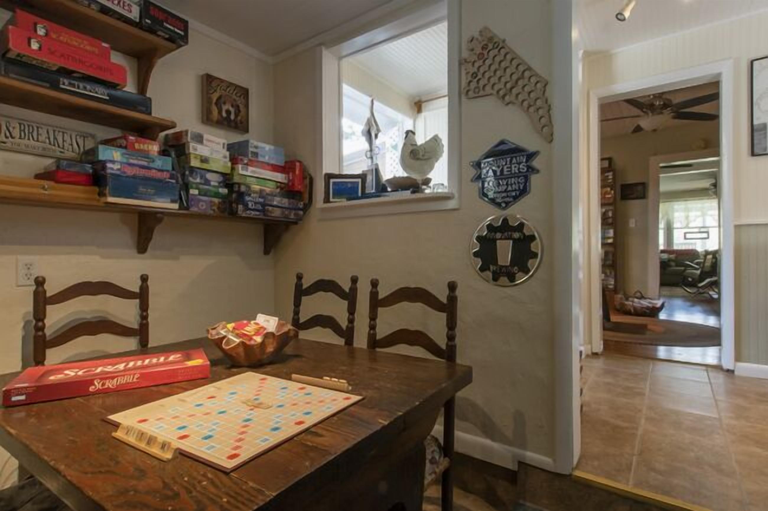 A dining area with a wooden table, chairs, and a Scrabble board. There are shelves with board games and a window with a view.