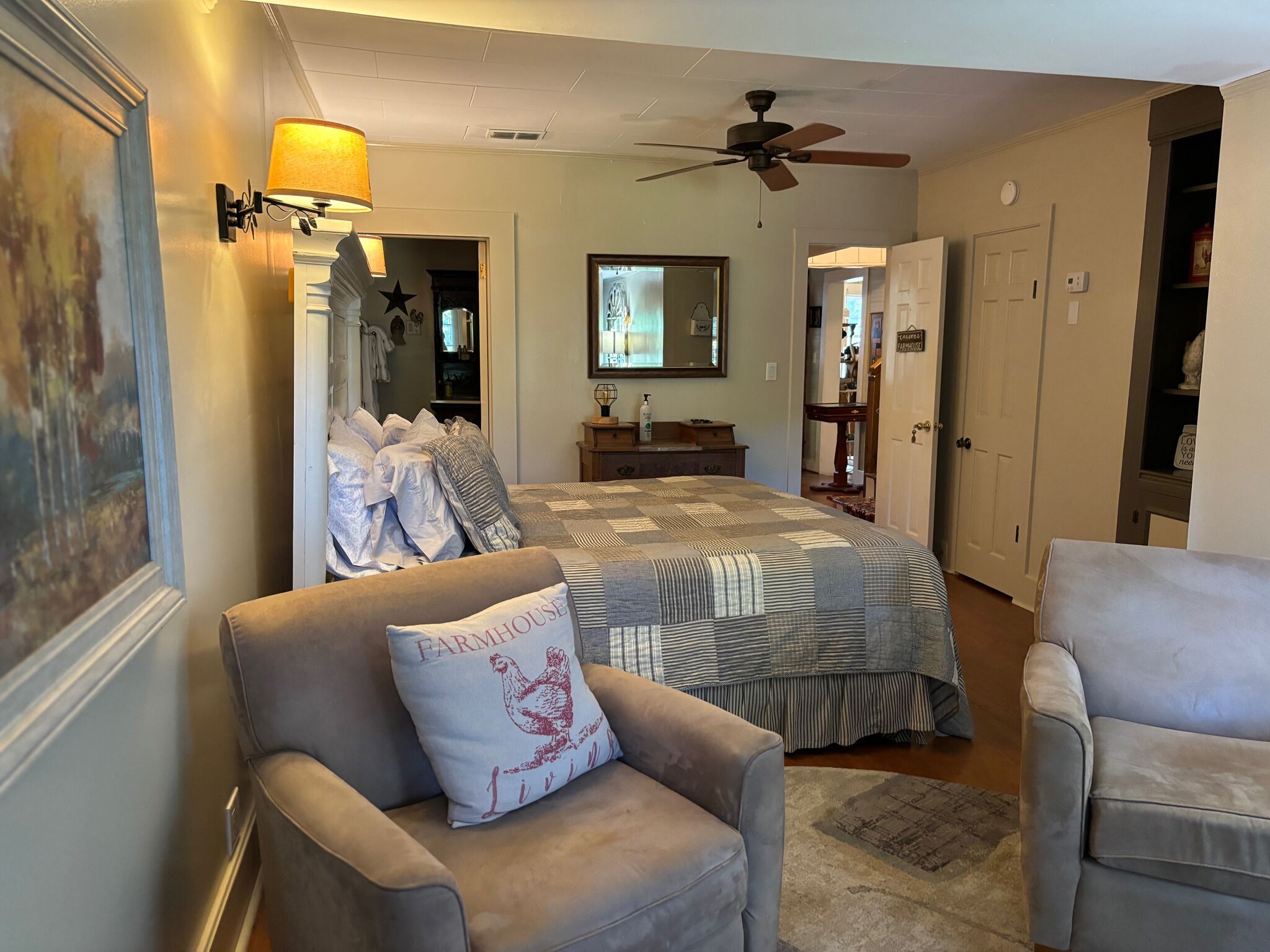 A bedroom with a bed, two armchairs, a ceiling fan, a lamp, and a door leading to another room.