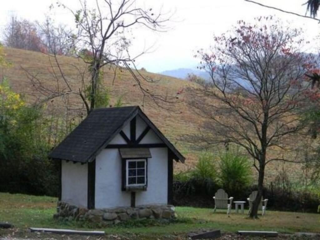 A small, white cottage with a black roof and stone foundation, surrounded by a grassy area and trees.