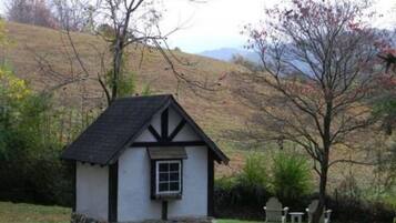 A small, white cottage with a black roof and stone foundation, surrounded by a grassy area and trees.