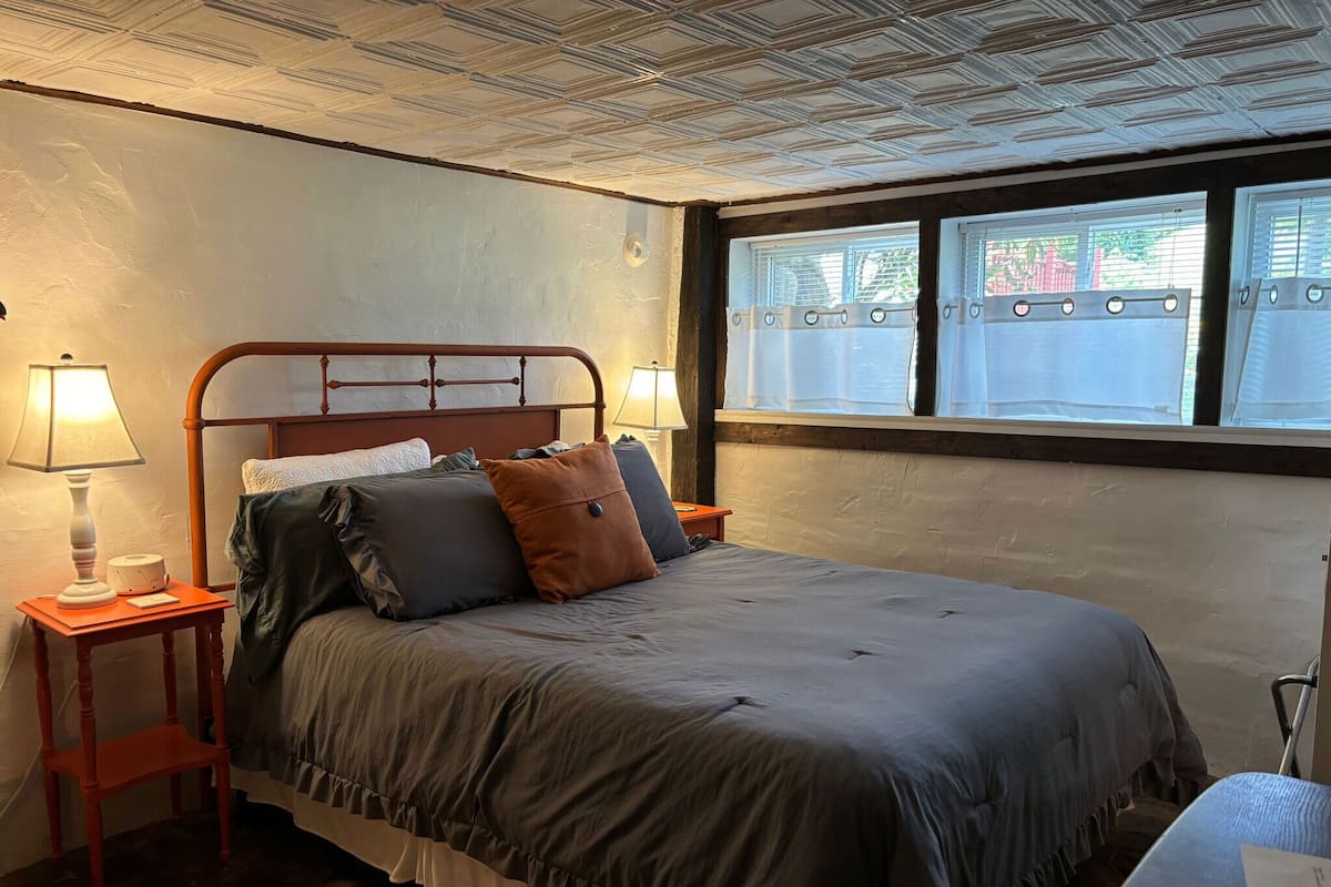 A bedroom with a bed, two bedside tables, a lamp, a window with blinds, and a patterned rug.