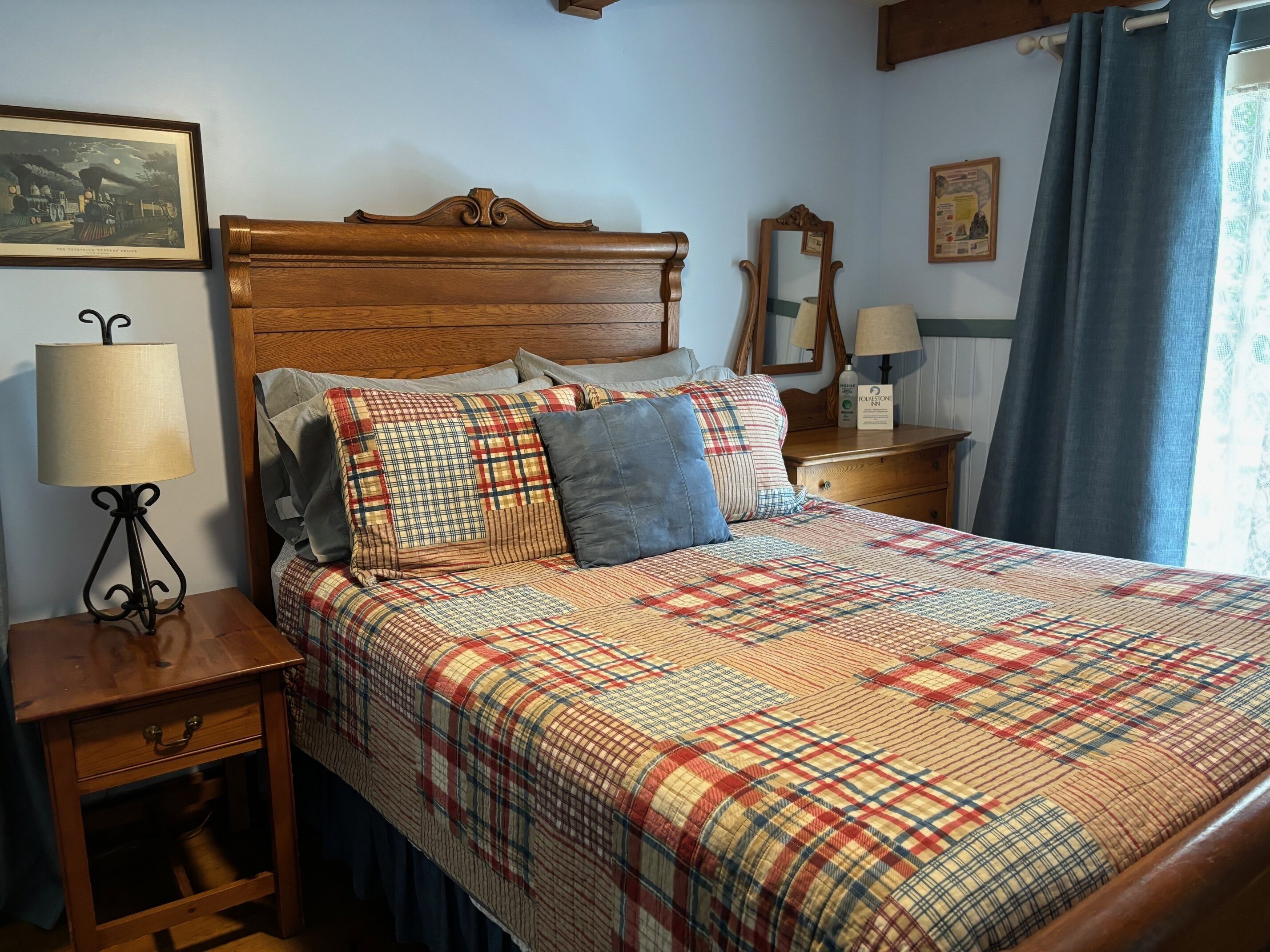 Un lit bien fait, avec une couette à carreaux, une tête de lit en bois et une table de chevet surmontée d’une lampe.